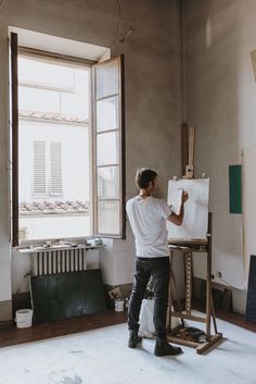 a man standing in front of an easel and drawing on a white board next to a window