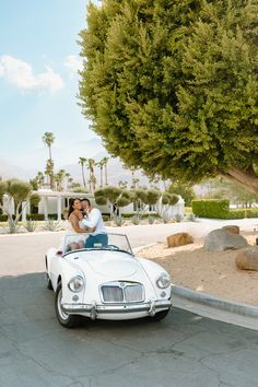 a man and woman sitting on top of a white car in front of a tree