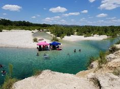 people are swimming in the clear blue water near some rocks and trees, while one person is holding a pink umbrella over his head