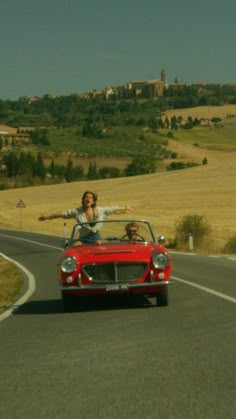 two people in a red convertible car driving down a road with hills and fields behind them