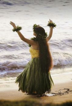 a woman in a grass skirt standing on the beach with her hands up to the sky