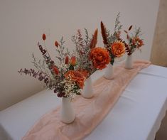 three white vases with orange flowers and greenery on a tableclothed cloth