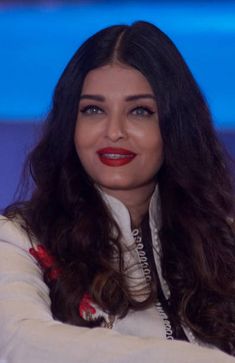 a woman with long dark hair wearing a white sweater and red lipstick sitting in front of a blue background