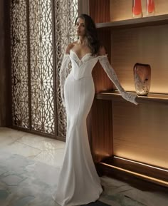 a woman in a white dress standing next to a shelf with vases on it