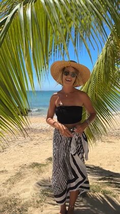 a woman standing under a palm tree on the beach wearing a straw hat and sunglasses