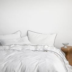 a bed with white linens and black piping on the pillows, along with a wooden night stand