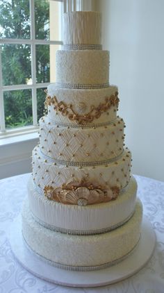 a white wedding cake with gold decorations on the top and bottom tier is sitting on a table in front of a window