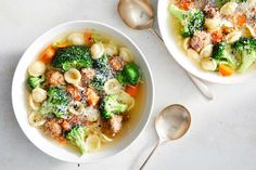 two bowls of pasta and broccoli soup with spoons on a white surface