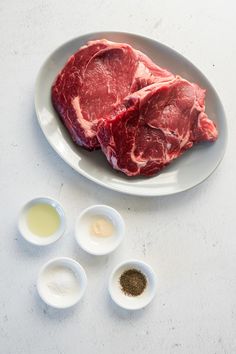 two pieces of raw meat on a plate with three small bowls and seasonings next to it