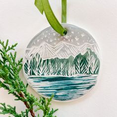 a christmas ornament hanging on a tree branch with water and mountains in the background