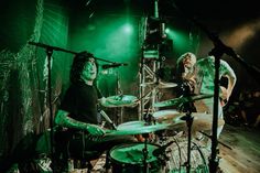 two men playing drums on stage with green lights in the background and one man standing behind them