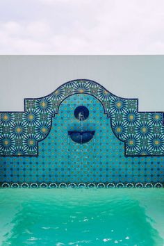 an artisticly designed fountain with blue and green tiles on the wall, in front of a pool