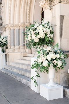 two tall white vases with flowers on the side of a building next to each other