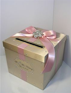 a gold box with pink ribbon and a diamond brooch on the top is shown