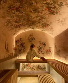 a woman sitting on top of a wooden bench in a room filled with floral wallpaper