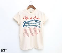 Set sail for style with this vintage-inspired Côte d'Azur graphic tee! Perfect for seafood lovers and travel enthusiasts, it captures the coastal charm and vibrant flavors of the French Riviera. The text on the bottom reads, "The Côte d'Azur, or French Riviera, is a glamorous Mediterranean destination known for its stunning beaches, luxurious resorts, and charming coastal towns. In addition to its breathtaking beauty, the region is famous for its fresh seafood, particularly sardines, sea bass, a French Beach Style, Italian Graphics, Vintage Aesthetic Retro, French Beach, Travel Tees, Aesthetic Retro, Retro Graphic Tees, Coastal Charm, Sea Bass