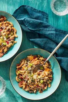 two bowls filled with pasta and vegetables on top of a blue table cloth next to silverware