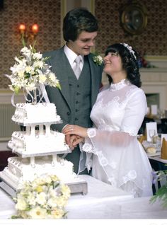 a bride and groom standing next to a wedding cake on a table with flowers in it