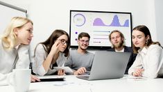 four people sitting at a table looking at a laptop screen with graphs on the wall behind them