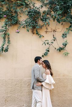 a man and woman standing next to each other in front of a wall covered with vines