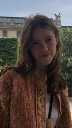 a woman standing in front of a building with trees and bushes behind her, smiling at the camera