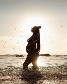 a pregnant woman standing in the ocean at sunset with splashing water all around her