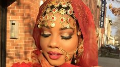 a woman wearing a red veil and headpiece with gold jewelry on her face standing in front of a brick building