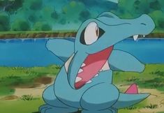 a blue dinosaur with its mouth open sitting on the ground next to a body of water