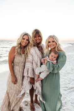 three women and a baby standing on the beach at sunset, smiling for the camera