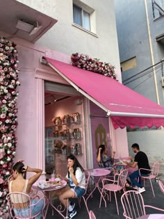 some people sitting at pink tables and chairs in front of a building with flowers on it