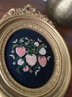 an ornate gold frame holds a cross - stitched bouquet of flowers in pink and green