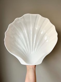 a person's hand holding up a large white shell shaped dish in front of a gray wall