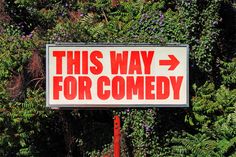 a sign that says this way for comedy in front of some trees and bushes with purple flowers