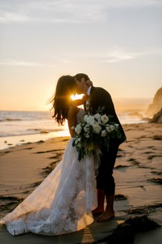 a bride and groom kissing on the beach at sunset