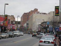 many cars are parked on the street in front of shops and businesses along with people walking down the sidewalk