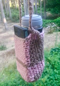 a crocheted cup holder hanging from a tree in the woods with a coffee mug on it