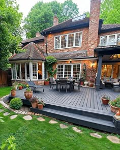 an outdoor deck with chairs and tables in front of a large brick house surrounded by greenery