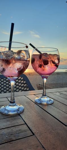 two glasses filled with liquid sitting on top of a wooden table next to the ocean