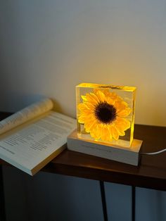 a yellow sunflower sitting on top of a table next to an open book and lamp