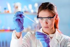 a woman in lab coat and goggles holding up a test tube with blue liquid