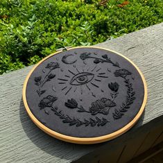 an embroidery project is shown on a wooden surface with plants and bushes in the background