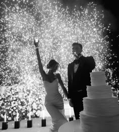 a bride and groom standing next to a wedding cake with fireworks in the background at night