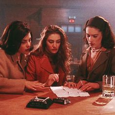 three women sitting at a table with papers and pens in front of them, talking