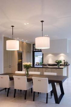 a dining room table with white chairs and lights hanging from it's ceiling in front of an open kitchen