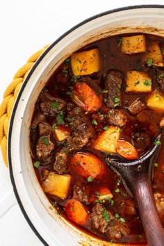 a pot filled with stew and vegetables on top of a white countertop next to a wooden spoon