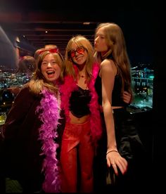 three women in costumes posing for the camera at a party with city lights behind them