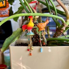 two colorful rooster earrings hanging from chains on a plant in front of a potted plant