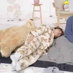 a man laying in bed next to a teddy bear and another person sleeping on the floor