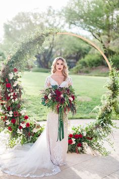 a woman standing in front of an arch with flowers and greenery on it, holding a bouquet