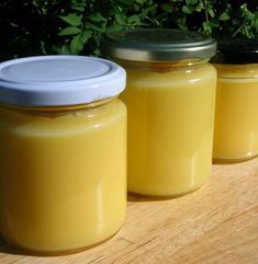 three jars filled with yellow liquid sitting on top of a wooden table next to bushes